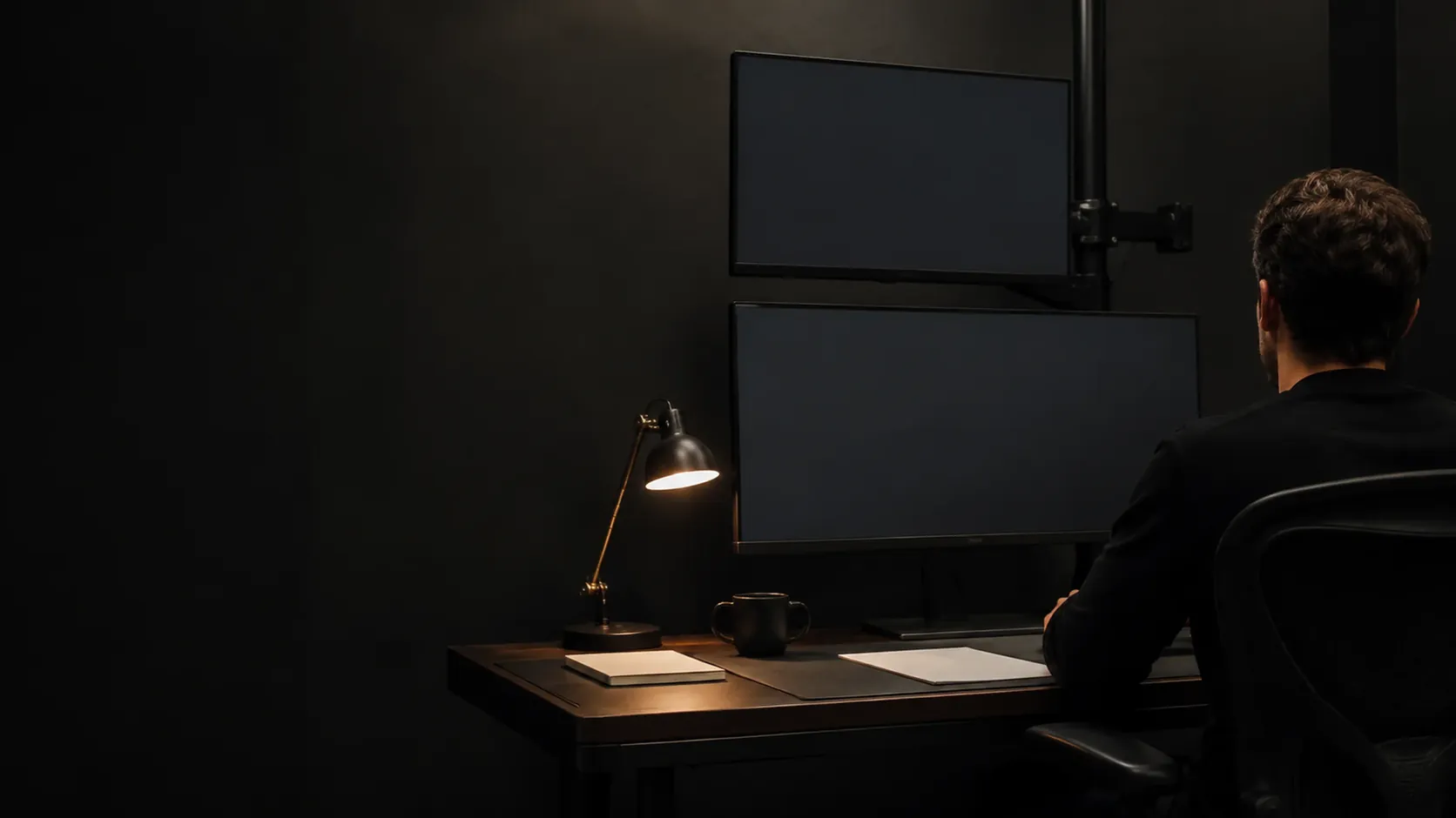 A person working late at two monitors in a dark office, one desk lamp lit beside them.